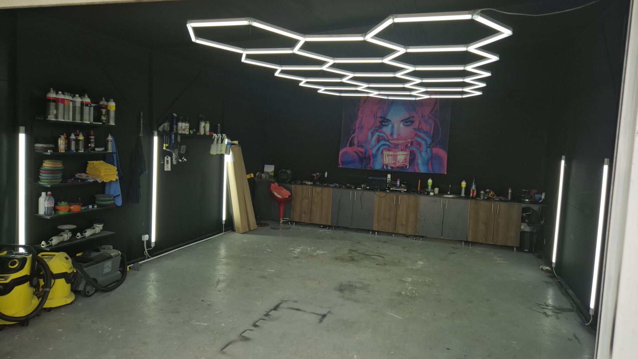 Interiorul atelierului: lampă hexagonală mare pe tavanul negru, raft cu produse de detailing și paduri de polish pe peretele din stânga, banc de lucru cu dulapuri și un tablou pe peretele din fund.