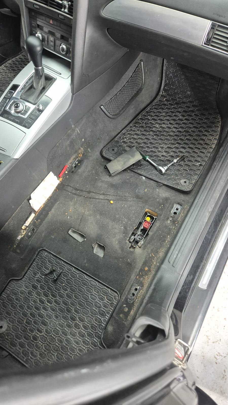 Podeaua din dreapta față a unui Audi A6, cu covorașele încă pe loc. Pe mochetă și pe prag se văd firimituri, praf și resturi mărunte, iar lângă scaun stau sculele cu care se demontează.