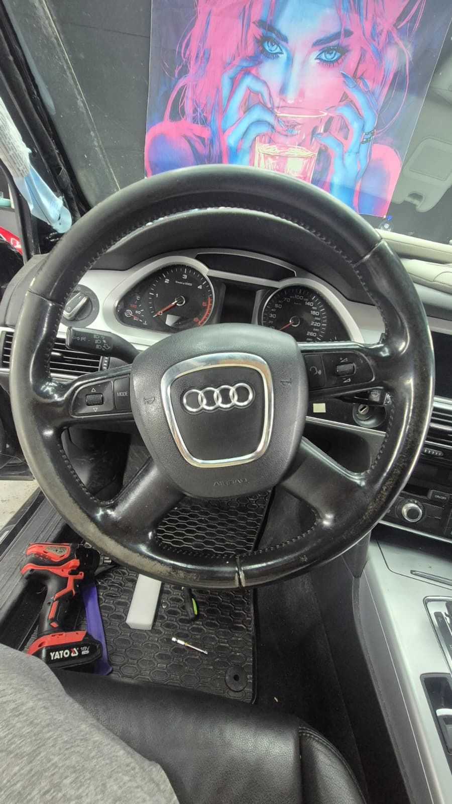 Volanul unui Audi A6, fotografiat din scaunul șoferului. Pielea de pe coroană e lucioasă și tocită în zonele unde stau mâinile, cu porțiuni decolorate spre gri, iar pe butoni s-a adunat depunere.