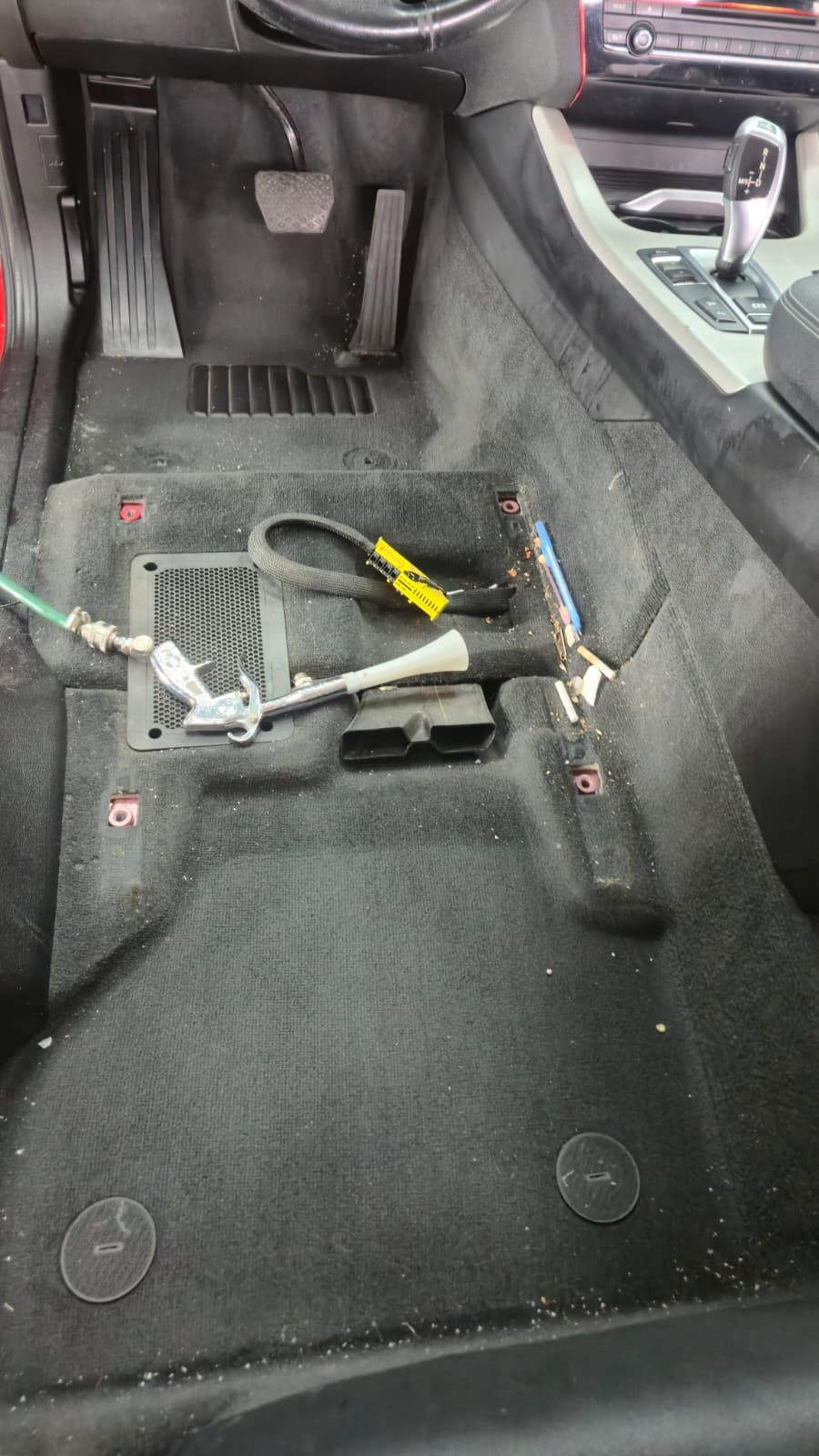Podeaua din stânga față a unui BMW X3, cu scaunul șoferului demontat. Pe mochetă se văd firimituri, un pix și resturi adunate sub scaun, plus furtunul aparatului de extracție.