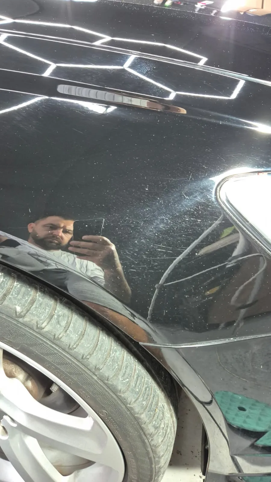 Aripa din față a unei mașini negre, cu roata în cadru. Pe lac se văd zgârieturi fine și stropi albi de apă uscată, iar în suprafață se oglindesc lampa din atelier și cel care fotografiază.