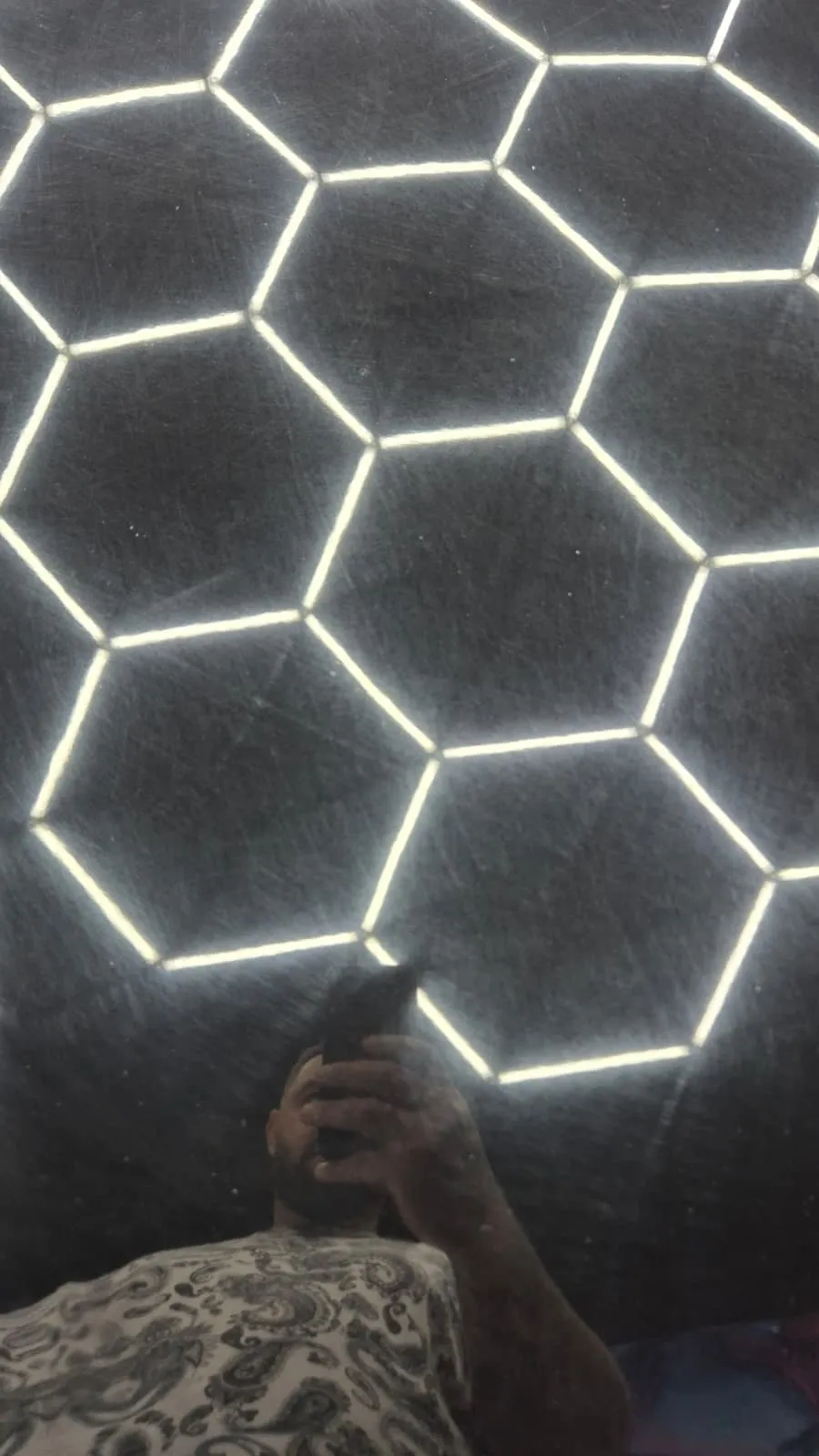 Prim-plan pe o suprafață neagră, sub lampa hexagonală din atelier. Hexagoanele se reflectă într-o rețea deasă de zgârieturi fine, care pornesc în toate direcțiile; jos se vede reflexia celui care fotografiază.