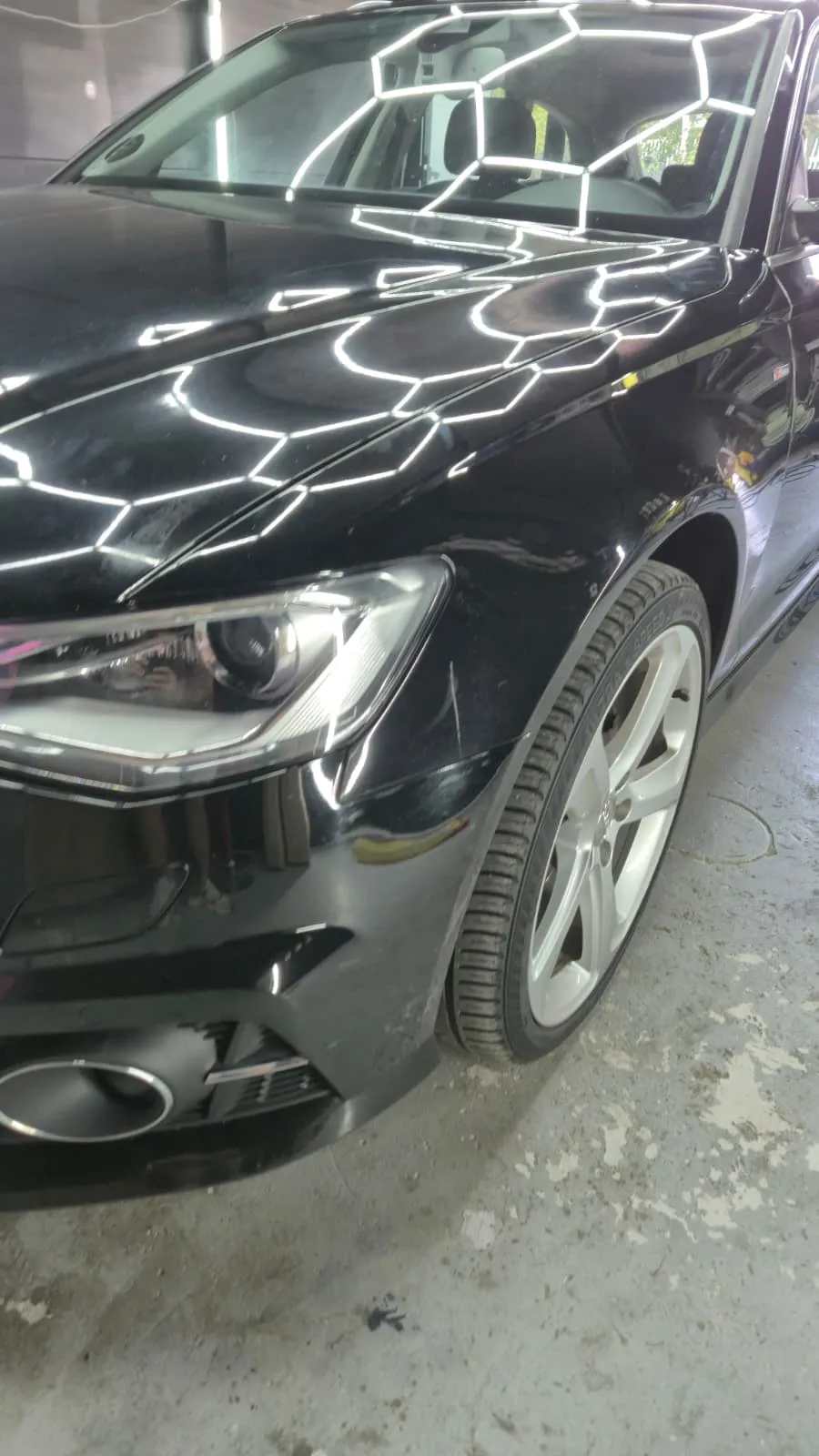 Partea din față a unei mașini negre sub aceeași lampă hexagonală: reflexia hexagoanelor are muchii drepte, fără haloul care apare pe un lac plin de zgârieturi.