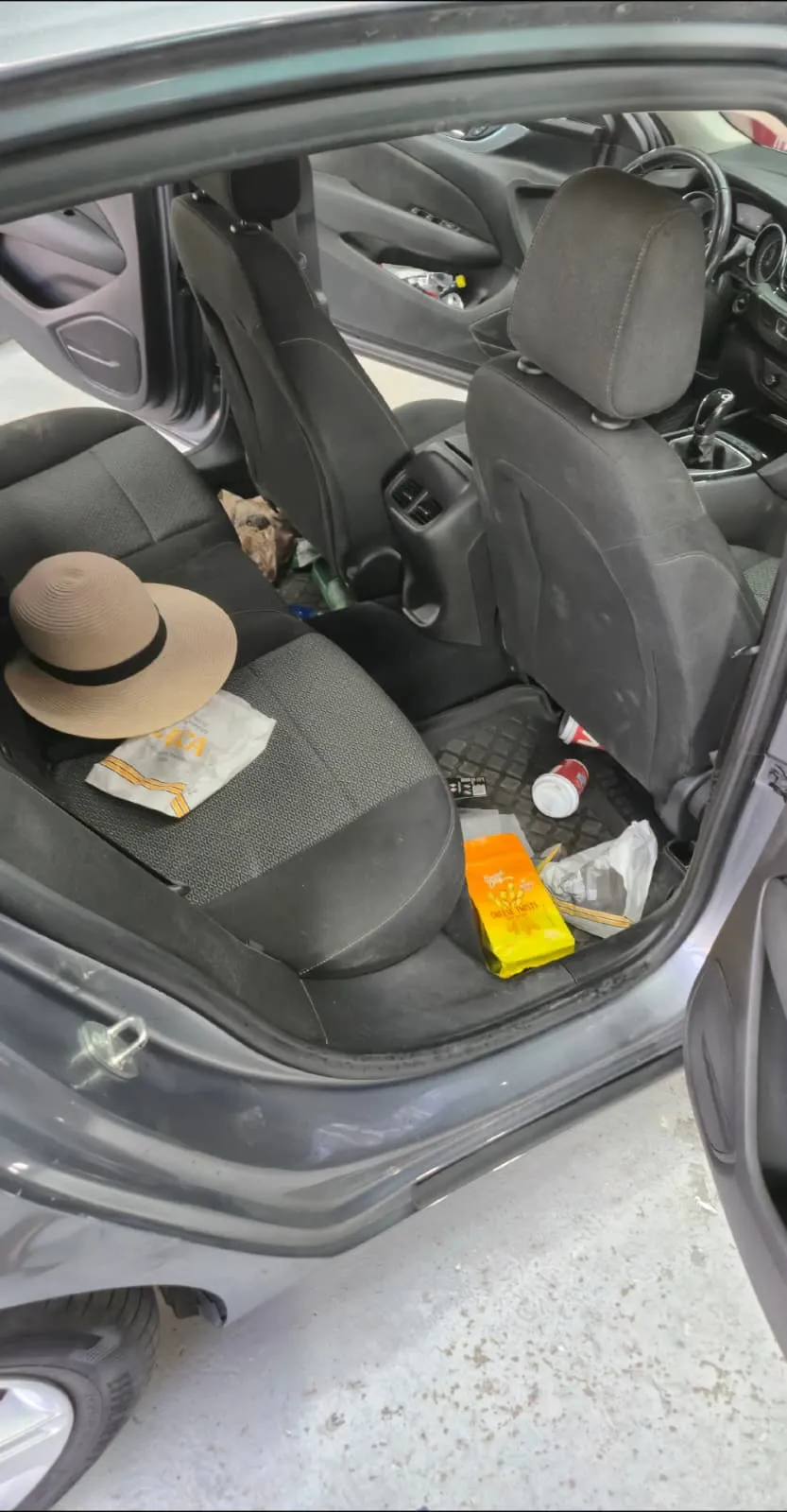 Bancheta din spate a unei mașini, fotografiată prin ușa deschisă, cu ambalaje, un pahar răsturnat și o pălărie lăsate pe scaun și pe podea, înainte de începerea lucrării.