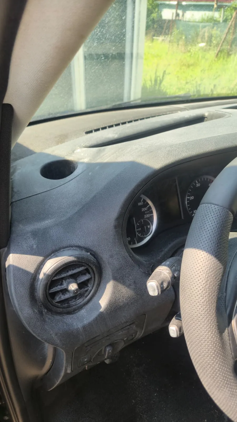 Prim-plan pe gura de ventilație din dreapta bordului, cu praf gri adunat pe lamele și pe rotița de reglaj, și un strat subțire de praf pe toată suprafața bordului din jur.
