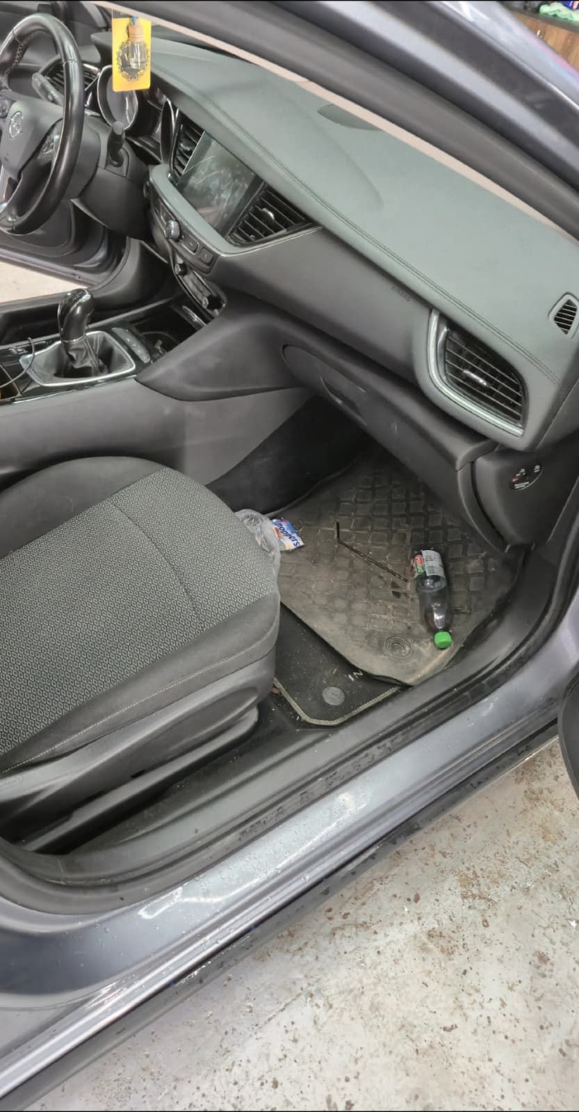 Podeaua din dreapta față a unui Opel Insignia: covoraș de cauciuc acoperit de praf, o sticlă goală și ambalaje căzute lângă prag, iar pragul are o dungă de murdărie prinsă în muchie.