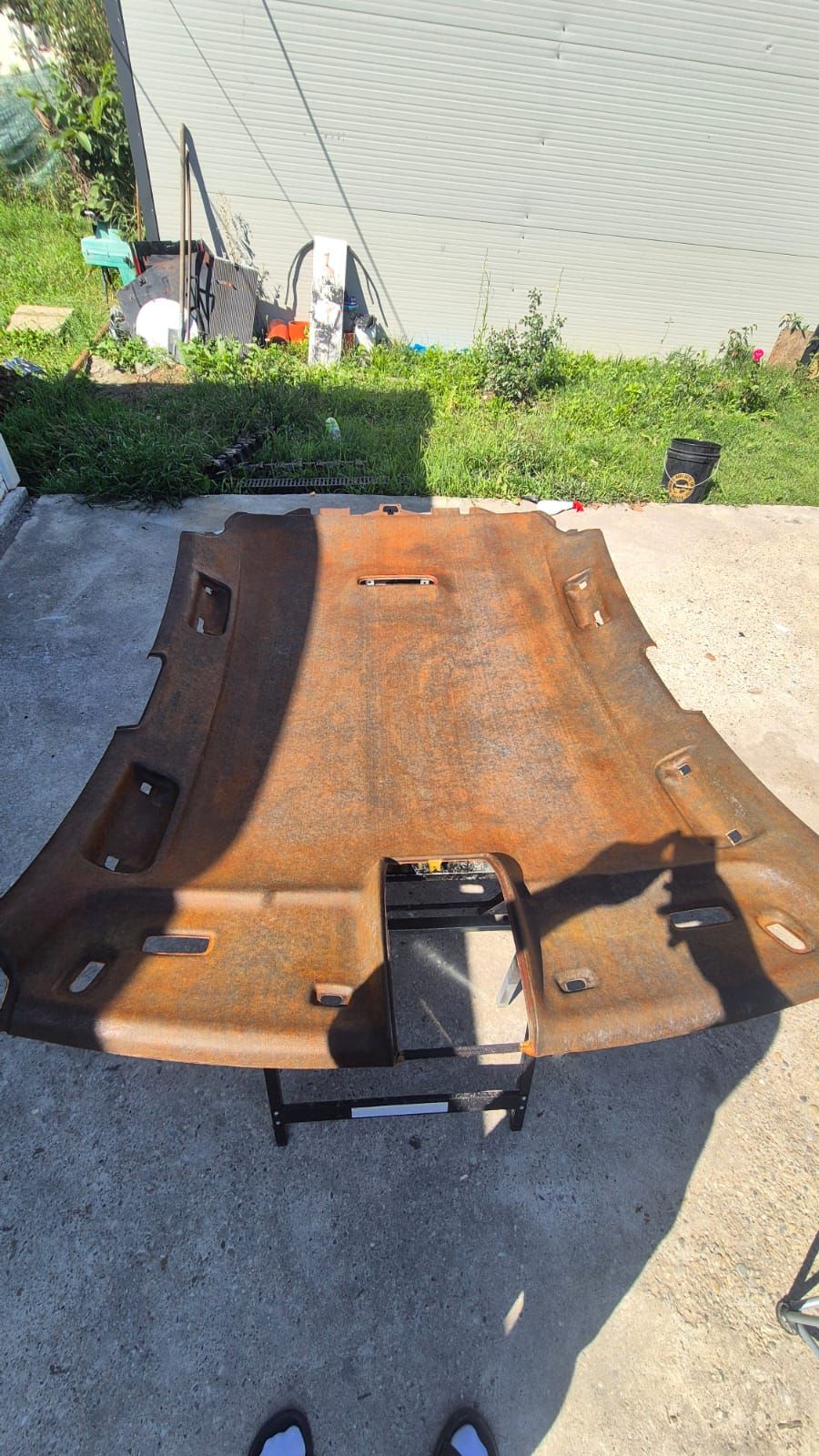 Plafonul demontat al unui Audi A6, sprijinit pe un suport în curtea atelierului, cu materialul vechi tras jos. Placa rămasă la vedere e pătată maro-ruginiu pe toată suprafața.